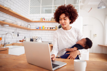 Wall Mural - Multi-Tasking Mother Holds Sleeping Baby Son And Works On Laptop Computer In Kitchen