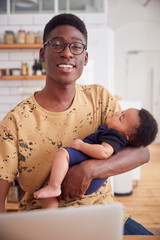 Portrait Of Multi-Tasking Father Holding Sleeping Baby Son And Working On Laptop Computer In Kitchen