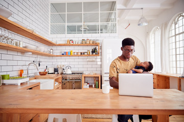 Wall Mural - Multi-Tasking Father Holds Sleeping Baby Son And Works On Laptop Computer In Kitchen