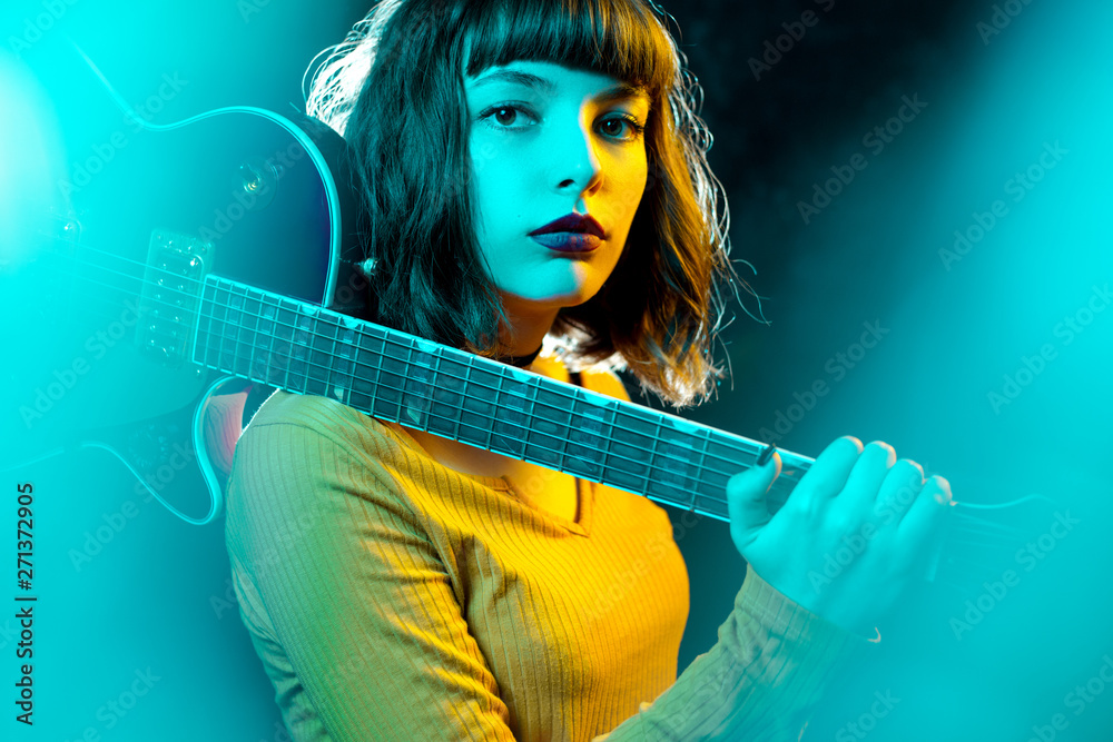 Beautiful young hipster woman with curly hair with red guitar in neon lights. Rock musician is playing electrical guitar. 90s style concept.