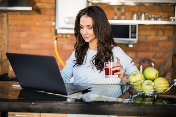 Young pretty woman drink juice using laptop computer in the kitchen with fruits.