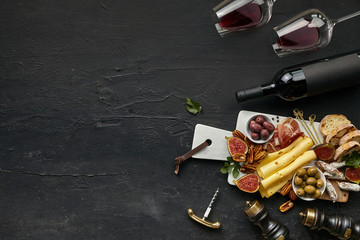 Two glasses and bottle of red wine and a tasty cheese plate with fruits, olives and toasted bread on a wooden kitchen plate on the black stone background, top view, copy space. Gourmet food and drink.