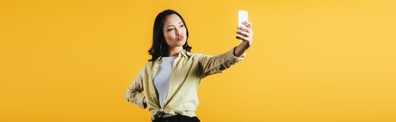 Wall Mural - brunette asian woman taking selfie on smartphone isolated on yellow