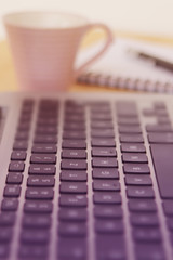 black laptop keyboard with selective focus and blur close-up, side view, a cup of coffee and a notebook at the back, soft rose tinting, vertical