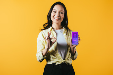 Wall Mural - smiling asian girl showing ok sign and smartphone with shopping app, isolated on yellow