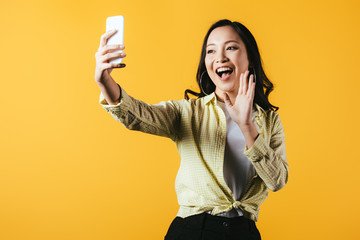 Wall Mural - excited asian girl waving and making video call on smartphone isolated on yellow