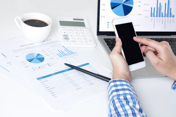 Poster - Asian businessmen use equipment for business. Holding a smartphone and viewing the screen with diagrams and financial information, And black screen mobile phones on modern white work desks.