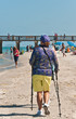Back view, of a seniior couple in sun protective clothes walking a tropical, sandy beach shoreline with walking poles, advancing on a wood pier with tourists, on a sunny, winter day on gulf of mexico