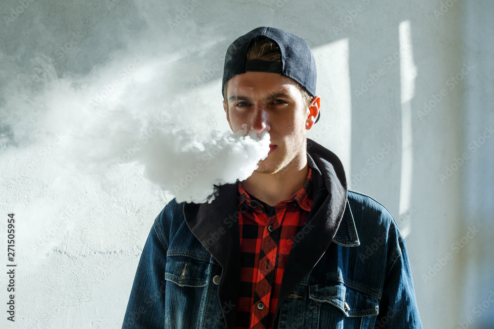 Vape teenager. Portrait of young handsome guy in a cap smoking an  electronic cigarette on the street in the spring. Bad habit that is harmful  to health. Vaping activity. Close up. Stock