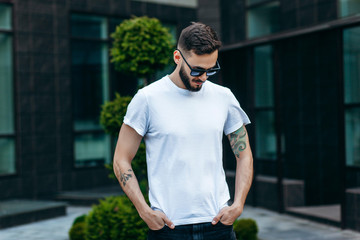 Wall Mural - A young stylish man with a beard in a white T-shirt and glasses. Street photo