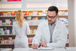 © bnenin - Focused pharmacist working at the counter, writing. Colleague working in the background.
