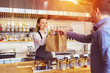 © DanRentea - Cheerful waitress wearing apron serving customer at counter in restaurant - Small business and service concept with young woman owner offering recycled paper bag with take away food to online client