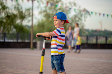 Two boys, one in a cap, the other in a hood, roll on a scooters pushing off a road with smiles on their face around square for walks and rest on  summer