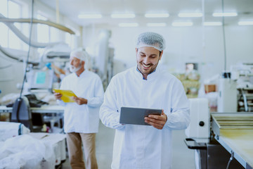 Young Caucasian smiling supervisor in sterile white uniform using tablet while standing in food plant. In background older one worker controlling machine.