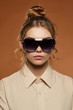 © RedUmbrella&Donkey - Cropped front shot of blonde lady, wearing shirt. The girl with bun and wavy hair locks is looking at camera against the brown background, wearing shield sunglasses with black rim and silver insert.