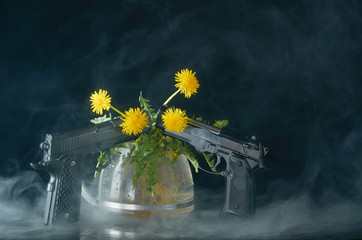 Dandelion with roots and leaves in a glass teapot and with two pistols on a dark background in smoke