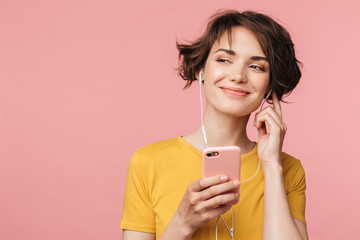 Wall Mural - Happy young beautiful woman posing isolated over pink wall background listening music with earphones using mobile phone.
