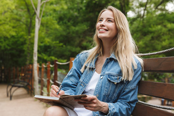 Wall Mural - Amazing happy young woman student posing outdoors in park writing notes in notebook.