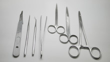 A Pair Of Stainless Steel Surgical Clamps, Scalpel Knife, Medical Grade Scissors, Forceps And Assorted Probes Arranged Straight On A White Table.