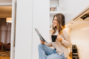 Wall Mural - Joyful dark-haired lady spending morning at home, reading newspaper with smile. Indoor photo of carefree girl in jeans enjoying tea in kitchen.