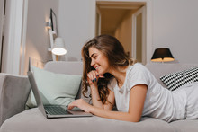 Amazing Curly Girl With Black Manicure Lying On Couch And Typing On Laptop Keyboard. Smiling Young Woman In White T-shirt Watching Movie On Computer, Chilling On Sofa.