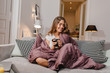 © Look! - Cheerful girl sitting on couch with blanket and cushions and smiling. Spectacular brunette lady laughing, while drinking coffee under plaid.