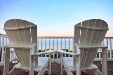 relax on Adirondack chairs in a desk balcony overlooking the beach and  ocean at sunset.  Add a beverage to this travel image.