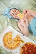 Cute girl with a towel on her head eats pizza in bed. Young woman eating pizza in bed. Life is a pleasure, body positive. Love for Italian food. Food habits, addiction to fast food.