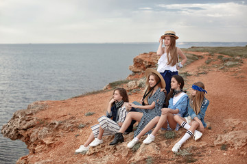 Five attractive and fun young girls having fun on the beach. Teenage sisters are resting together on the rocky shore of the blue ocean on a cloudy day against the blue sky and the coastline.