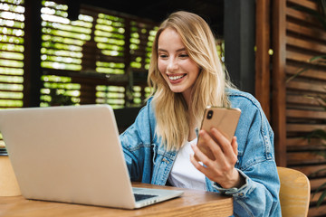 Wall Mural - Excited happy young woman posing outdoors in cafe using laptop computer and mobile phone.