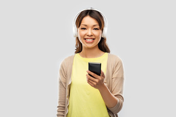 people, technology and audio equipment concept - happy asian young woman in headphones listening to music on smartphone over grey background