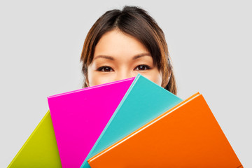 Poster - education, school and knowledge concept - asian woman or student girl hiding behind notebooks over grey background