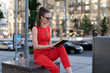 © Денис Бухлаев - A young European girl in a red suit and glasses is sitting on the parapet near the sidewalk and the roadway and reading a book, looking at a notebook. Copy space.