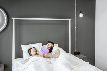 Wall Mural - Man reading book for a woman while lying together in the bed before the sleep at home