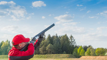Trap Or Skeet Shooting, Man In Red Clothes  Shoots From A Shotgun At Clay Pigeon,  A Background Of Forest And Sky, Images With Copy Space