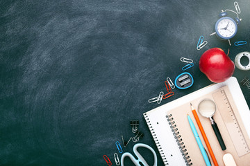 School stationary on chalkboard. Notebooks, pens, pencils, alarm clock, red apple and other tools. Flat lay with place for text.