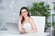 © Yarkovoy - A young brunette girl with glasses in a white dress is sitting in the office at the table. Woman works at the laptop. Low vision or fashion accessory