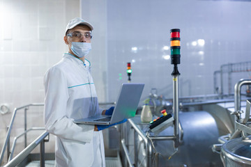 Wall Mural - Portrait of man in a white robe and a cap standing in production department of dairy factory with laptop
