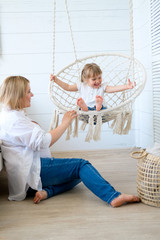 Beautiful little girl swinging in a hanging chair in the bedroom with her mom. A woman is pregnant with her second child. Macrame wicker chair, knitted blanket, straw pillow. Cozy home, happy family