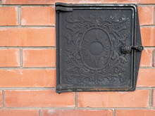 Russian Stove. Metal Black Door With Patterns On The Brick Red Wall.