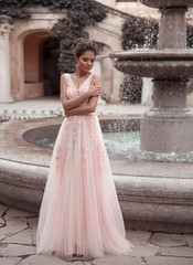 Beautiful bride in pink wedding dress. Outdoor romantic portrait of attractive brunette woman with hairstyle in prom dress with tulle skirt posing by fountain at park.