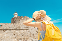 Happy Tourist Woman On Vacation Posing With Hat And Backpack In Front Of The Old Ruined Fort And Lighthouse In Rhodes, Greece