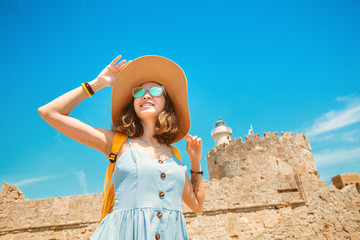 Wall Mural - Happy tourist woman on vacation posing with hat and backpack in front of the old ruined fort and lighthouse in Rhodes, Greece