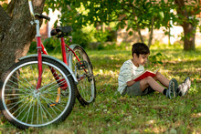 The Boy Sits On The Grass In The Garden And Thinks About The Book Read.  A Red Bicycle Stands By A Tree, A Book Is Lying On The Grass