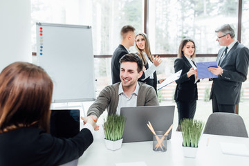 Wall Mural - We have a deal! Men shaking hands  woman and looking at each other with smile while sitting at the business meeting with their coworkers in modern office..