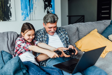 Grandpa and granddaughter holding joysticks and playing games on modern laptop.. Lying on modern house.