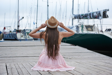 Brown-haired woman with long hair sits on a wooden pier, looks at the beautiful yachts