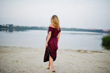 Wall Mural - Back of blonde sensual barefoot woman in red marsala dress posing against lake.