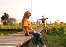 Asian Tanned Woman Wearing A Pretty Yellow Top Sitting On A Wooden Walkway Flower Plantation - Young Thai Female Smiling And Happy With Bright Sunshine In A Sunflower Farmers Field With Scarecrows
