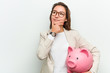 © Asier - Young european business woman holding a piggy bank looking sideways with doubtful and skeptical expression.